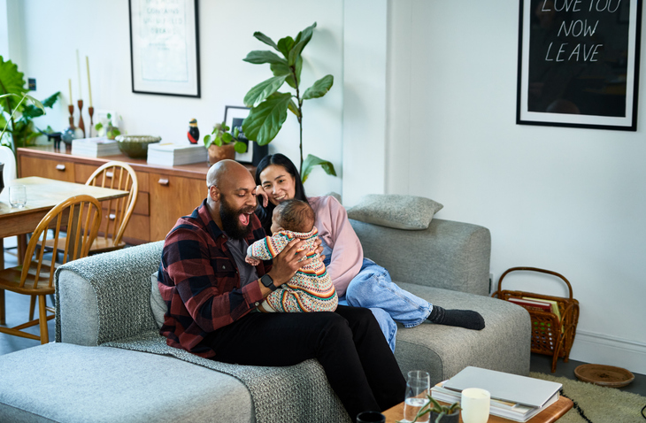 Multiracial parents sitting on sofa with baby son on black mid adult father’s lap and Chinese mature mother smiling
